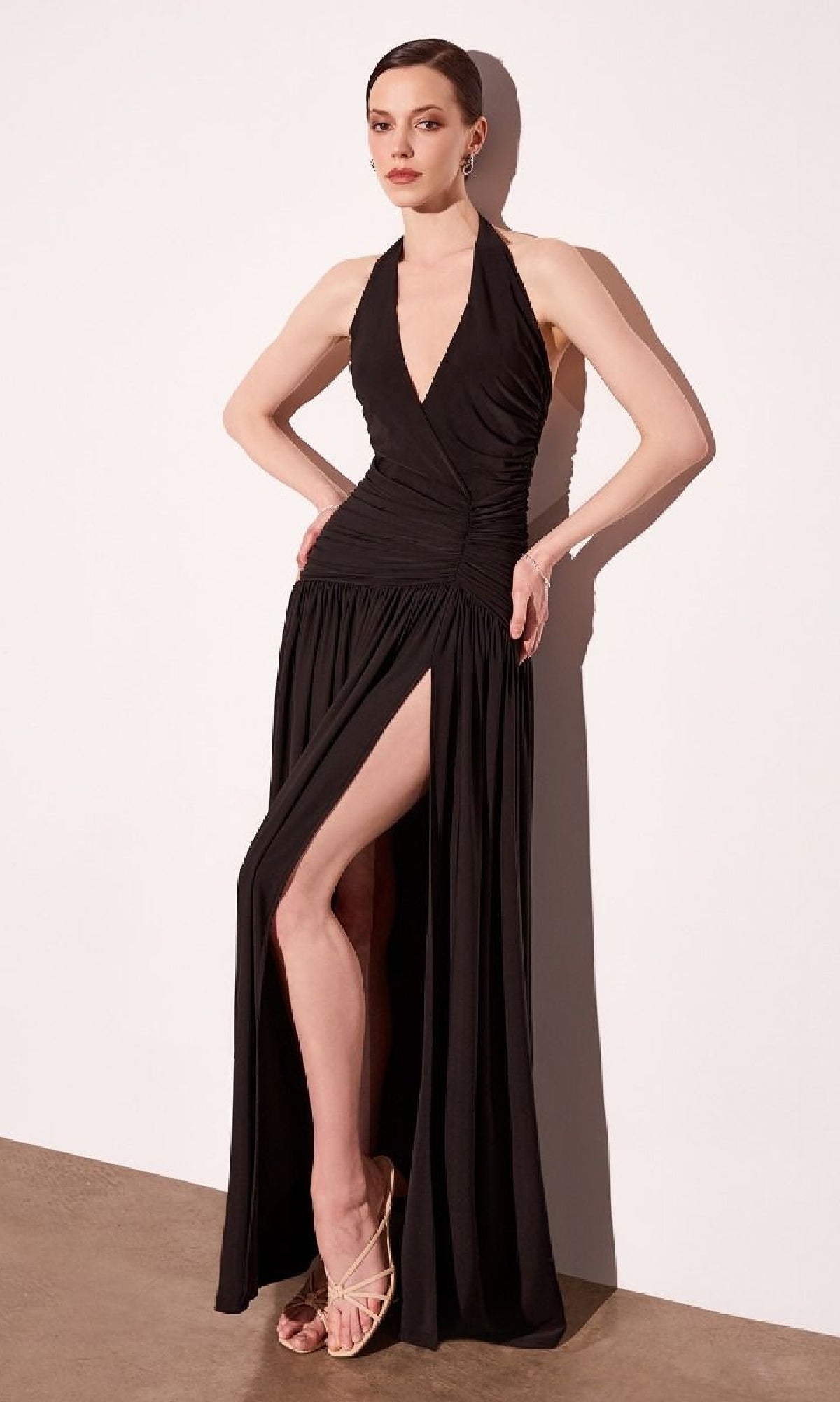A woman stands against a white wall wearing the Amoris T502 Backless Halter Prom Dress with a high slit and deep V-neck, paired with strappy beige heels.